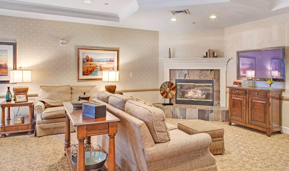 A cozy living room with beige patterned carpet and wallpaper, featuring a beige upholstered sofa and armchair, a wooden side table with a lamp and decorative items, a wooden console table, a fireplace with a tiled surround, and a flat-screen TV mounted on the wall above a wooden cabinet.