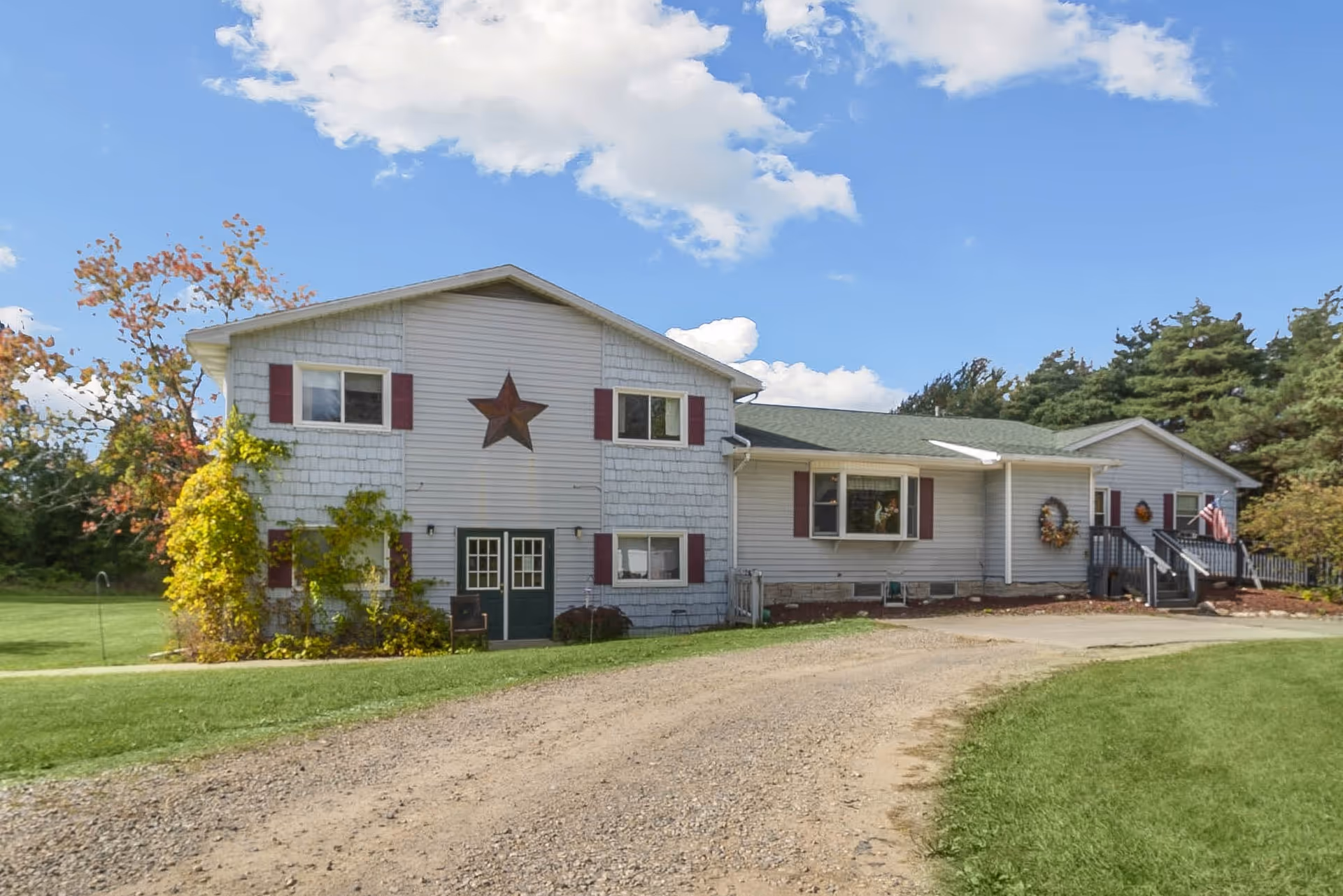 Exterior view of a single-story building with light gray siding and maroon shutters, featuring a large star decoration on the front. There is a gravel driveway leading up to the building, surrounded by green grass and trees under a partly cloudy blue sky.