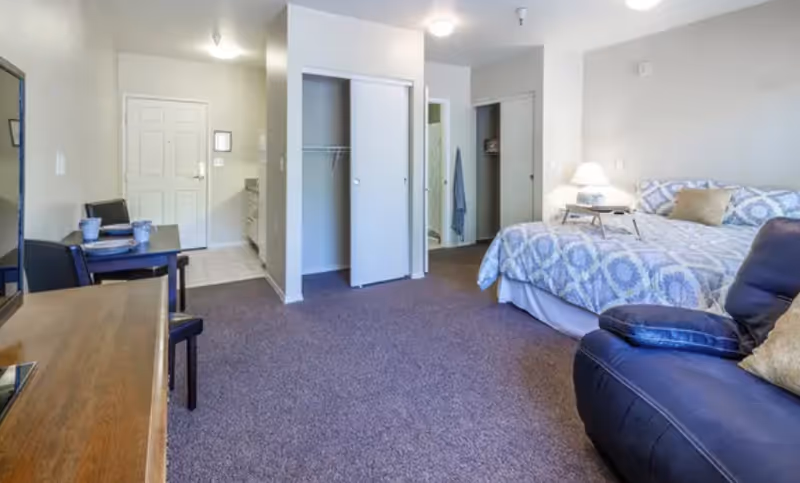A senior living facility room featuring a bed with patterned bedding, a bedside table with a lamp, a recliner chair, an open closet with sliding doors, a small dining table with two chairs, and a dresser with a mirror. The room has carpeted flooring and neutral-colored walls.