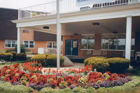 Entrance of a brick building with a covered porch supported by white columns. In front of the entrance is a colorful flower bed with red, purple, and white flowers, along with neatly trimmed bushes. There are chairs on the porch and a flagpole in the center of the flower bed.