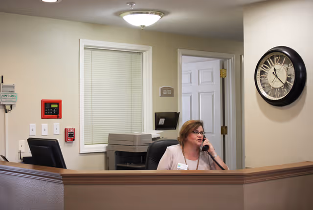 Reception area in a senior living facility with a woman sitting behind the desk talking on the phone. The desk has a computer monitor and a printer behind it. A large wall clock shows the time as 12:05. There is a door and a window with closed blinds behind the receptionist.