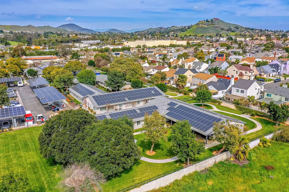 Aerial view of a residential facility with multiple buildings featuring solar panels on the roofs, surrounded by green lawns, trees, and a walking path. In the background, there is a neighborhood with houses and hills under a partly cloudy sky.