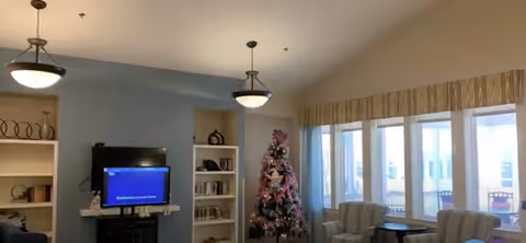 A cozy common room with a TV and bookshelves, seating by large windows, pendant lights, and a decorated Christmas tree.