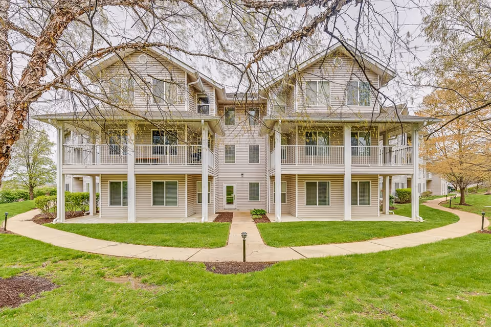 Exterior view of a two-story residential building with beige siding and white railings on the balconies. The building is surrounded by green grass, trees with budding leaves, and curved concrete walkways leading to the entrances.