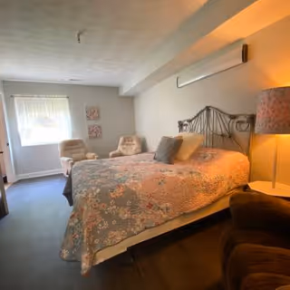 A cozy bedroom with a bed covered in a floral quilt, two beige armchairs near a window with sheer curtains, a bedside table with a lamp, and a cushioned chair in the foreground. The walls are light-colored, and there are two small decorative wall hangings near the window.