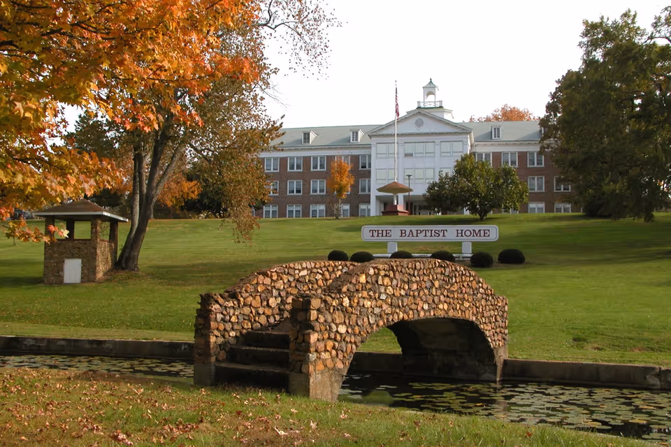 A large brick building with white trim and multiple windows sits atop a grassy hill with autumn trees around. In front of the building is a stone arch bridge over a small waterway with lily pads. A sign near the building reads 'THE BAPTIST HOME'.