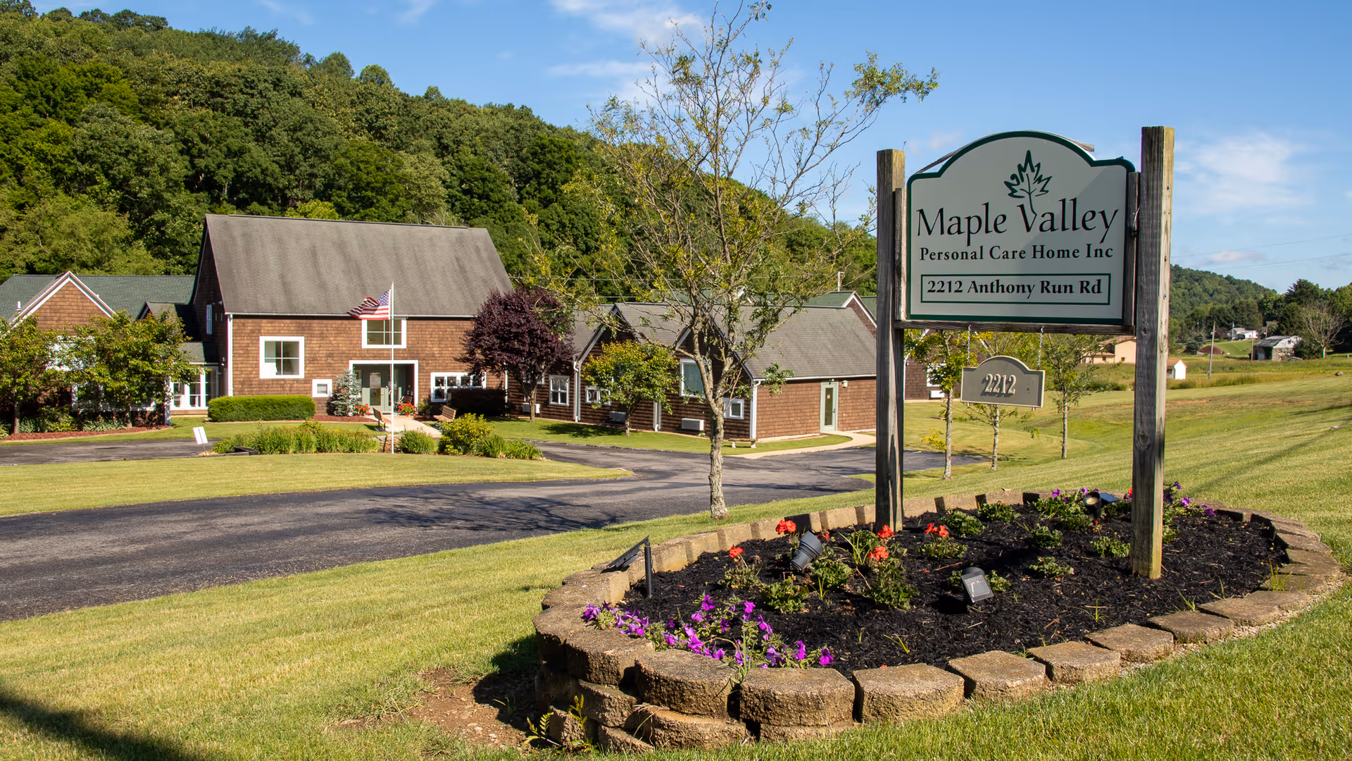 Exterior view of Maple Valley Personal Care Home showing a large sign with the facility's name and address in front of a landscaped flower bed. Behind the sign, there is a paved driveway leading to a brown building with white trim and an American flag in front. The area is surrounded by green grass and trees under a clear blue sky.