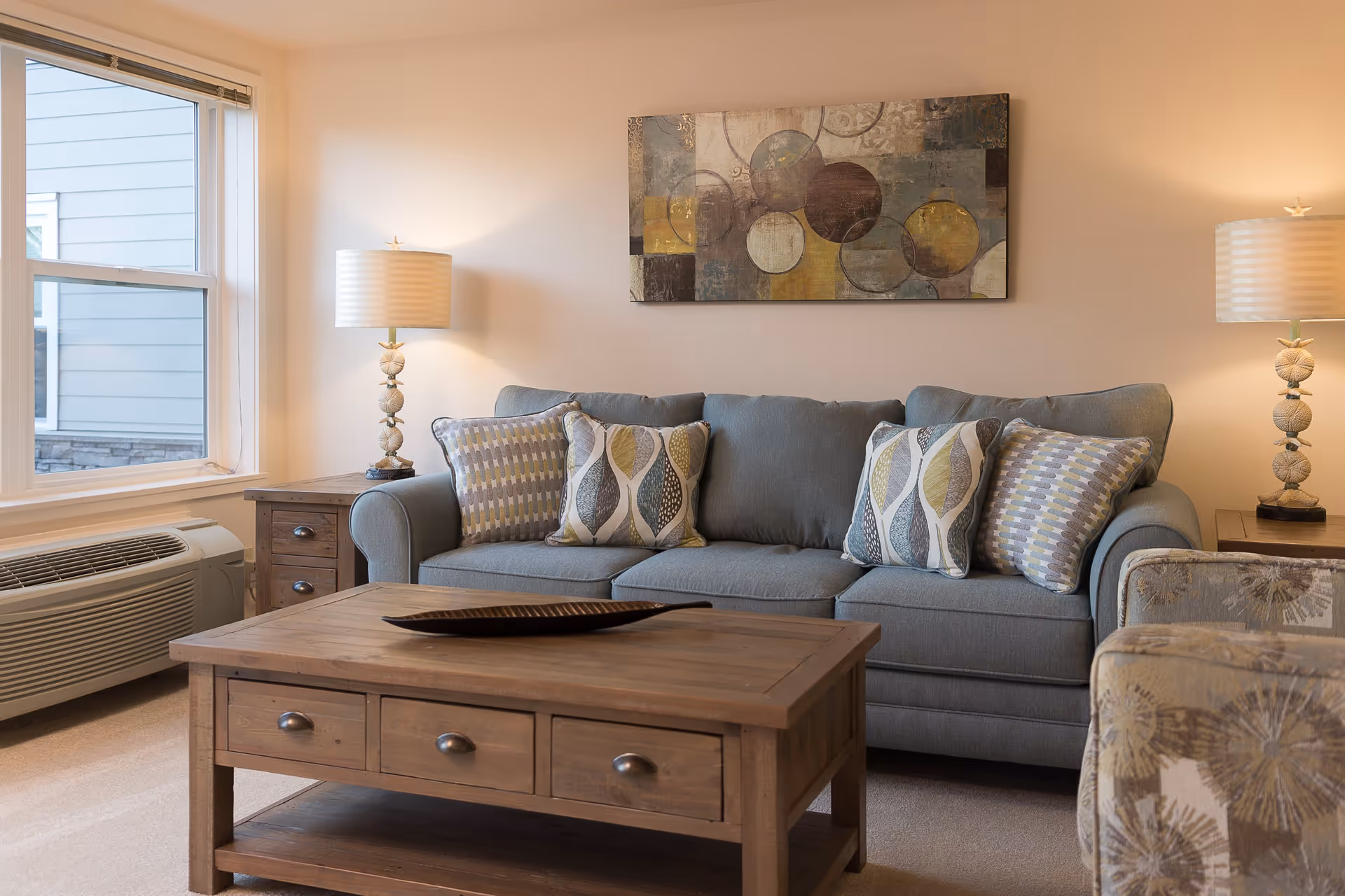 A cozy living room with a gray sofa adorned with patterned cushions, two matching table lamps on wooden side tables, a wooden coffee table with drawers, a large window, and a modern abstract painting on the wall.