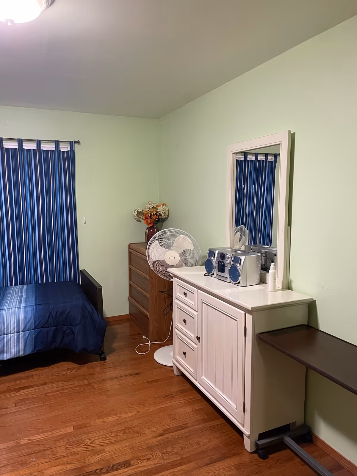 A small bedroom with a single blue bed, blue-striped curtains, a white dresser with mirror and stereo, a wooden chest, and a standing fan on hardwood floors.