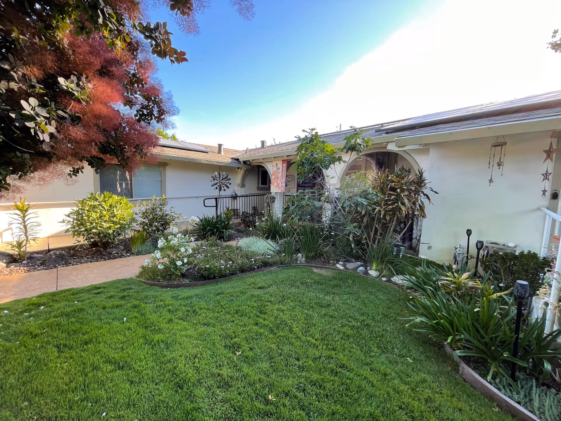A well-maintained garden area with green grass, various plants, and shrubs in front of a single-story building with a white exterior. The building has a covered entrance with an archway, decorative hanging stars, and a flag. The sky is clear and blue.