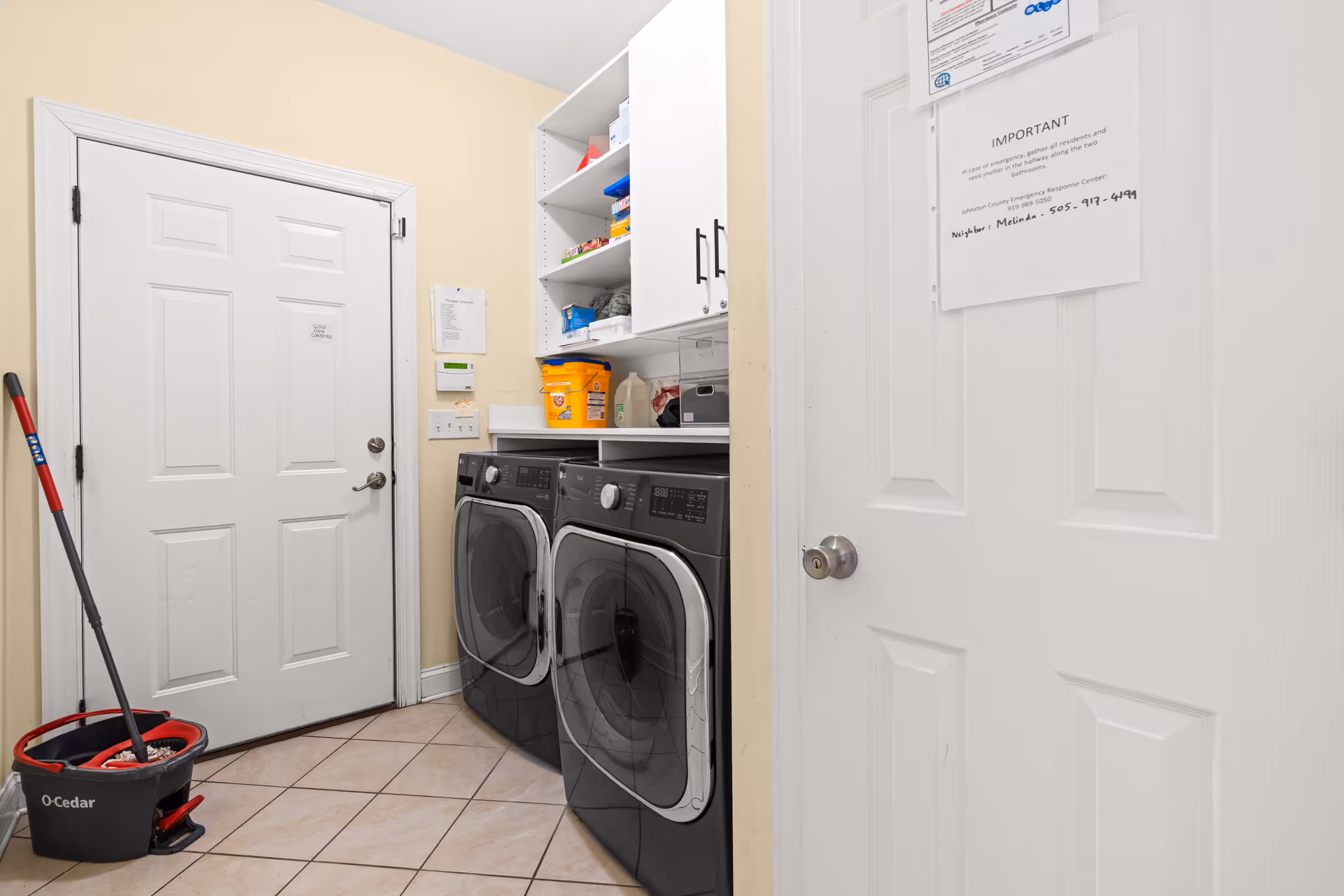 Small laundry room with front-loading washer and dryer, shelving stocked with supplies, and a mop bucket by the door.