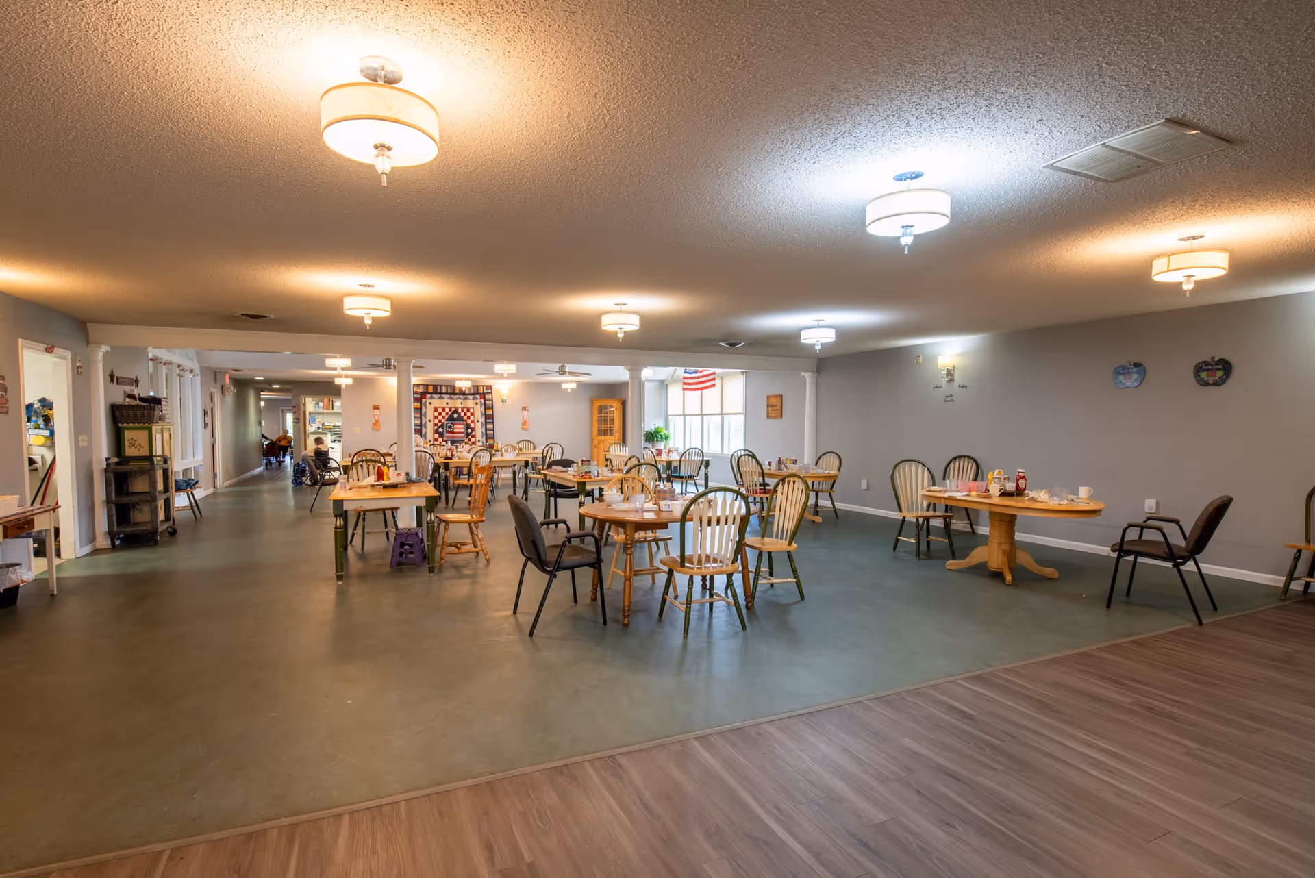 A spacious dining room in Wynwood House with multiple wooden tables and chairs arranged throughout the room. The room is well-lit with ceiling lights and has a mix of green and wood flooring. There is a quilt hanging on the far wall, an American flag near a window, and various decorations on the walls. A hallway is visible in the background with a person in a wheelchair.
