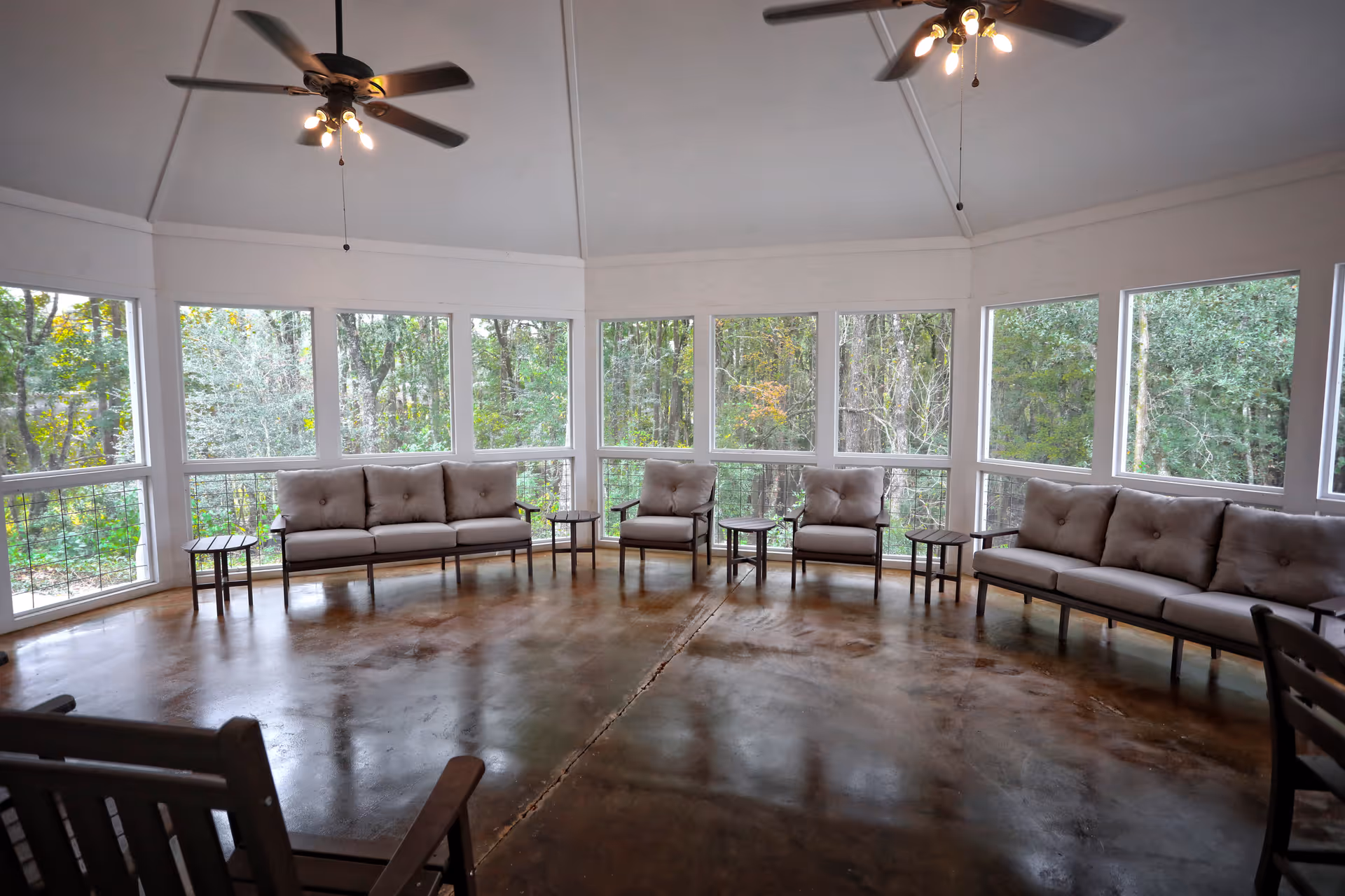 Spacious sunroom with cushioned sofas and chairs arranged around the perimeter, ceiling fans overhead, and large windows overlooking trees.