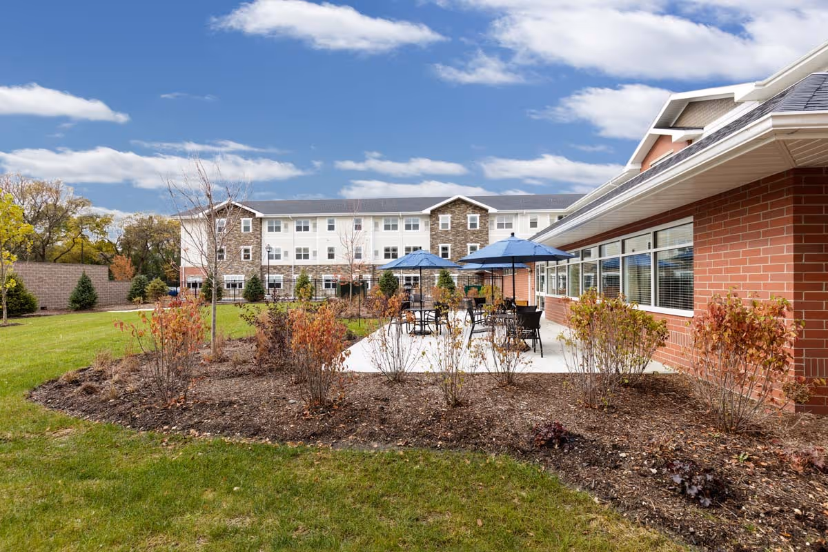 Outdoor patio with tables and blue umbrellas in front of a multi-story senior living building surrounded by landscaping.