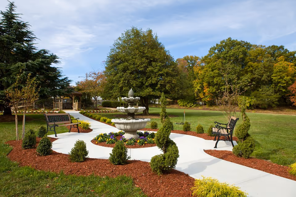 A landscaped outdoor garden area with a white concrete pathway winding around a tiered stone fountain surrounded by colorful flowers. There are two black metal benches on either side of the path, small trimmed bushes, and large trees in the background under a partly cloudy sky.