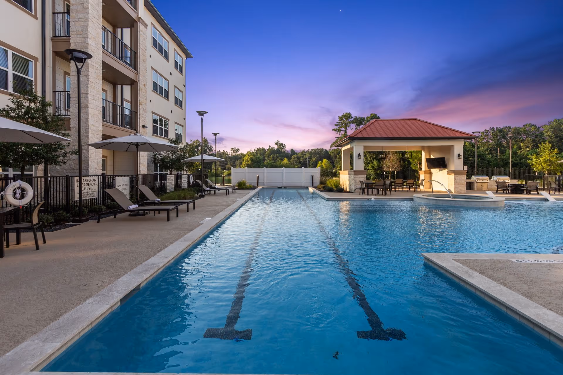 Outdoor swimming pool at a senior living facility during sunset, with lounge chairs and umbrellas along the poolside, a covered seating area with tables and chairs, and a building with balconies on the left side.