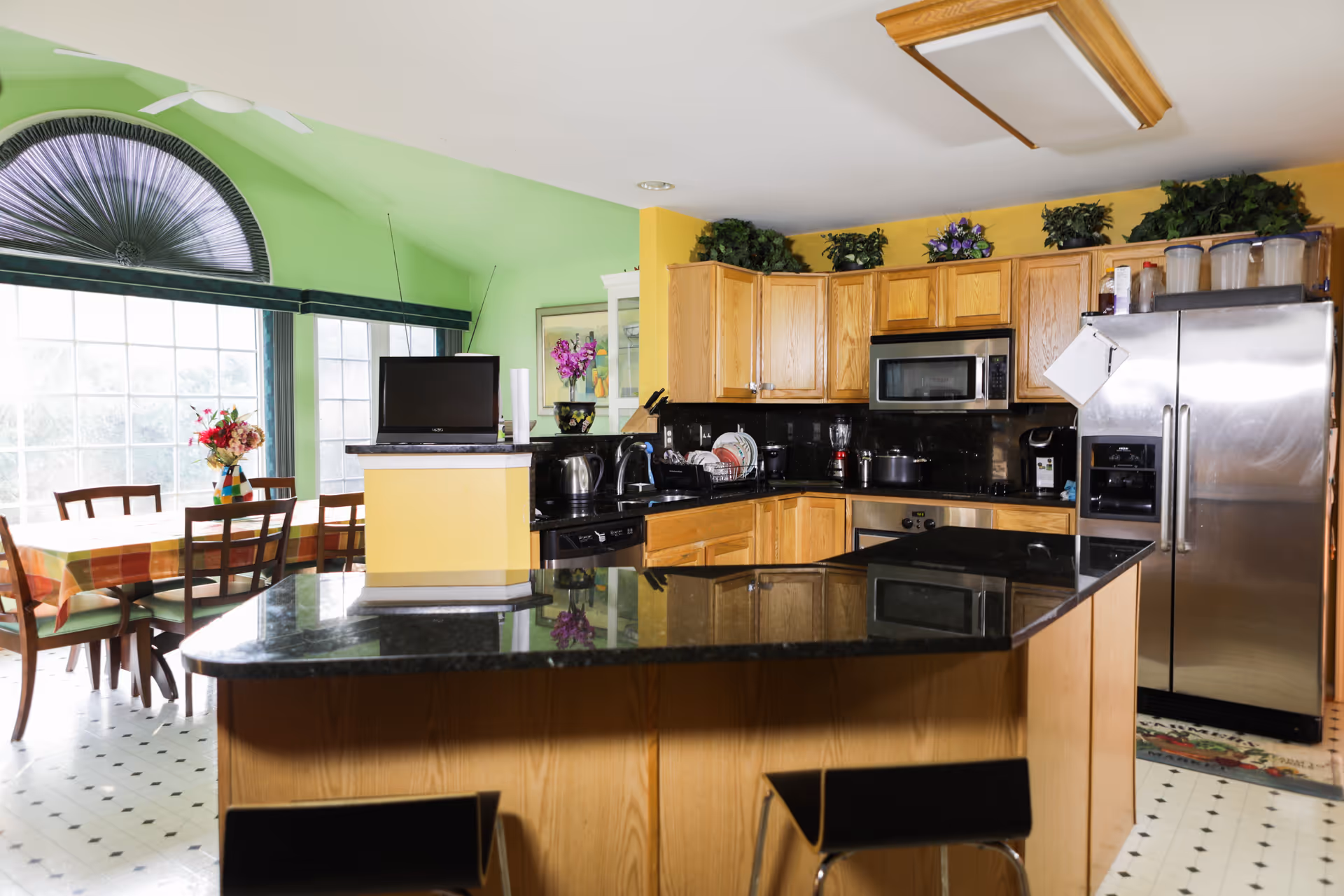 Bright kitchen with wooden cabinets, black granite countertops, stainless steel refrigerator, microwave, and various kitchen appliances. Adjacent dining area with a table covered by a checkered tablecloth and wooden chairs, large windows with green walls and a decorative fan-shaped window treatment.