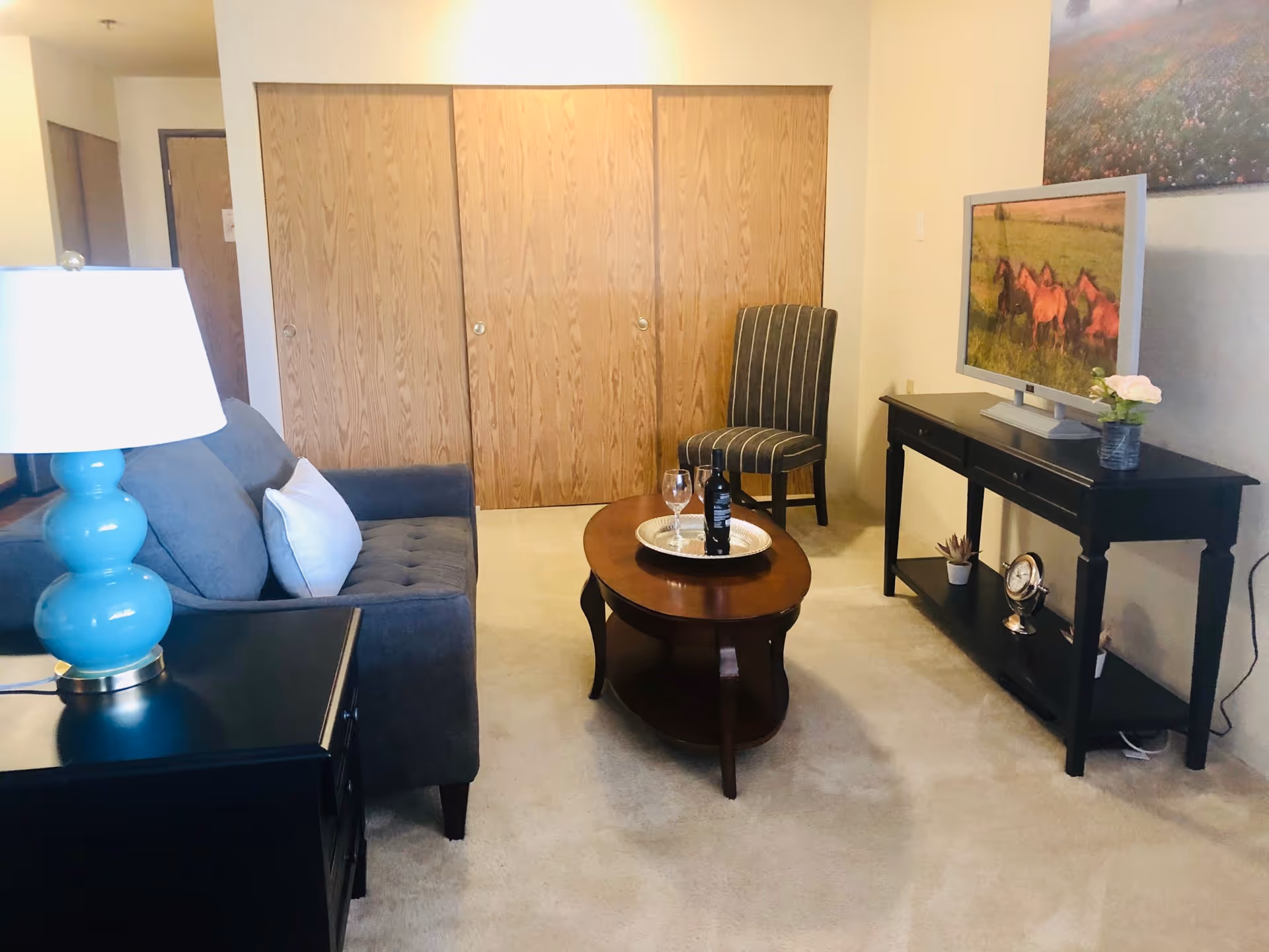 A cozy living room with a gray sofa, a blue lamp on a black side table, a wooden coffee table with a bottle of wine and two glasses, a striped chair, and a black TV stand with a flat-screen TV displaying an image of horses in a field. There are wooden closet doors in the background and a large wall art above the TV.