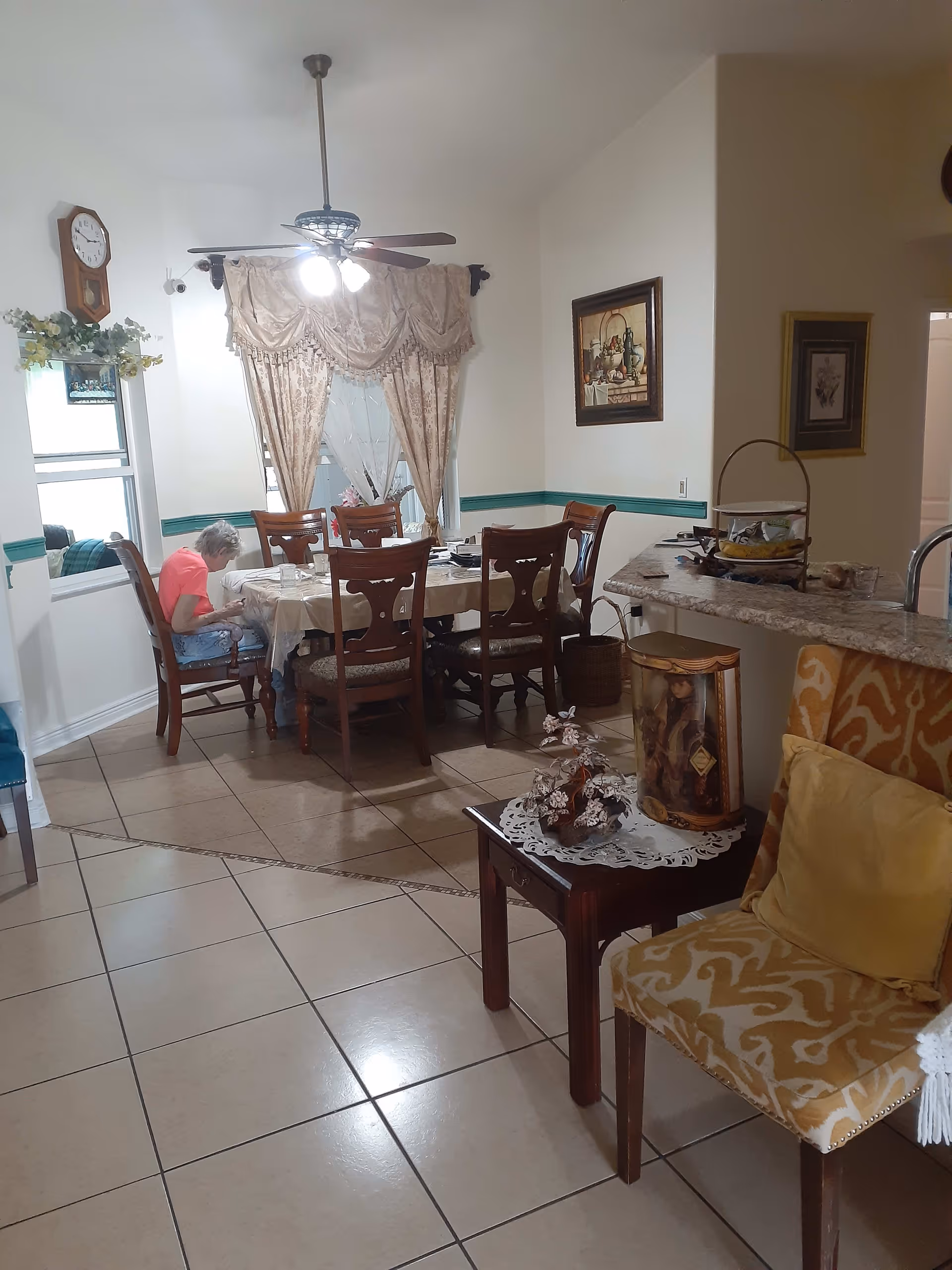 Interior view of a dining area in a senior living facility with a wooden dining table and six chairs. A ceiling fan with lights hangs above the table. There is a person sitting at the table wearing a pink shirt. The room has tiled floors, beige walls with a green trim, and decorative curtains on the window. A small side table with a floral arrangement and a yellow patterned chair with a cushion are visible in the foreground. The kitchen counter with snacks and a framed picture on the wall are also visible.