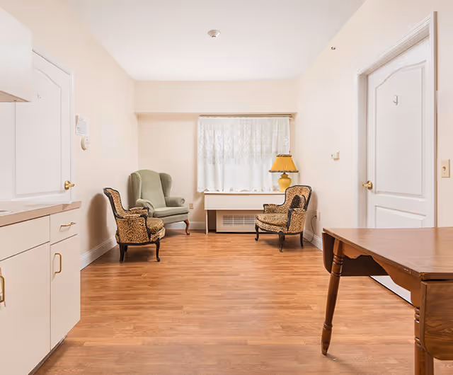 Bright, sparsely furnished living room with three armchairs, a wooden table, a lamp, and a window with lace curtains.