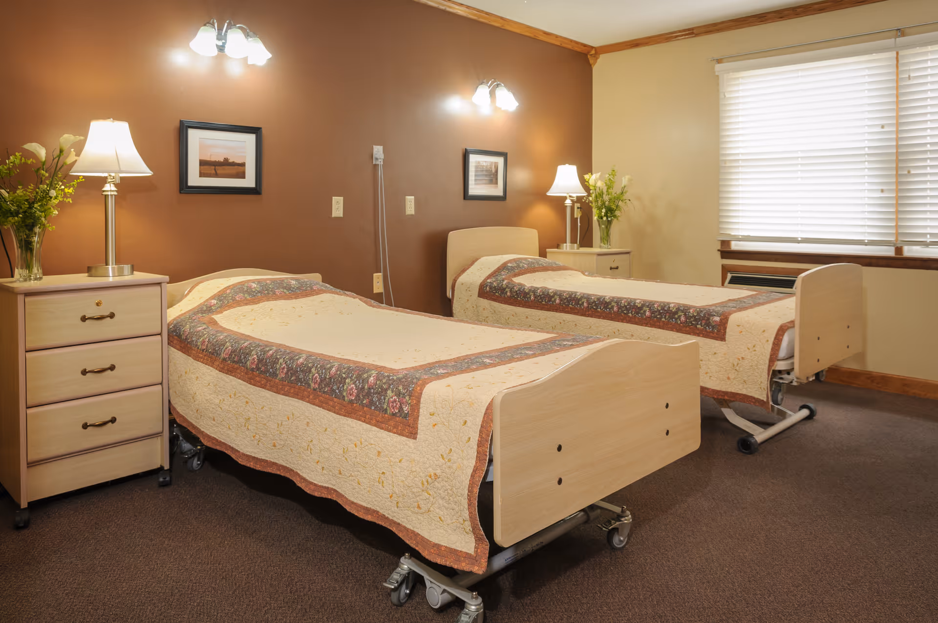 A bedroom with two hospital-style beds, each covered with a beige quilt featuring a floral border. Each bed has a wooden headboard and footboard. Between the beds is a small nightstand with a lamp and a vase of flowers. The walls are painted brown and beige, with framed pictures hanging above the beds. A window with closed blinds is on the right side of the room.