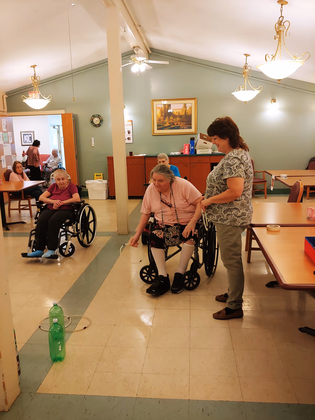 A group of elderly women, some in wheelchairs, are engaged in an indoor activity in a common room with tables and chairs. One woman in a wheelchair is reaching toward two green bottles placed on the floor. Another woman stands beside her, smiling and assisting. The room has light green walls, ceiling fans, and hanging lights, with a framed picture on the wall and cabinets in the background.