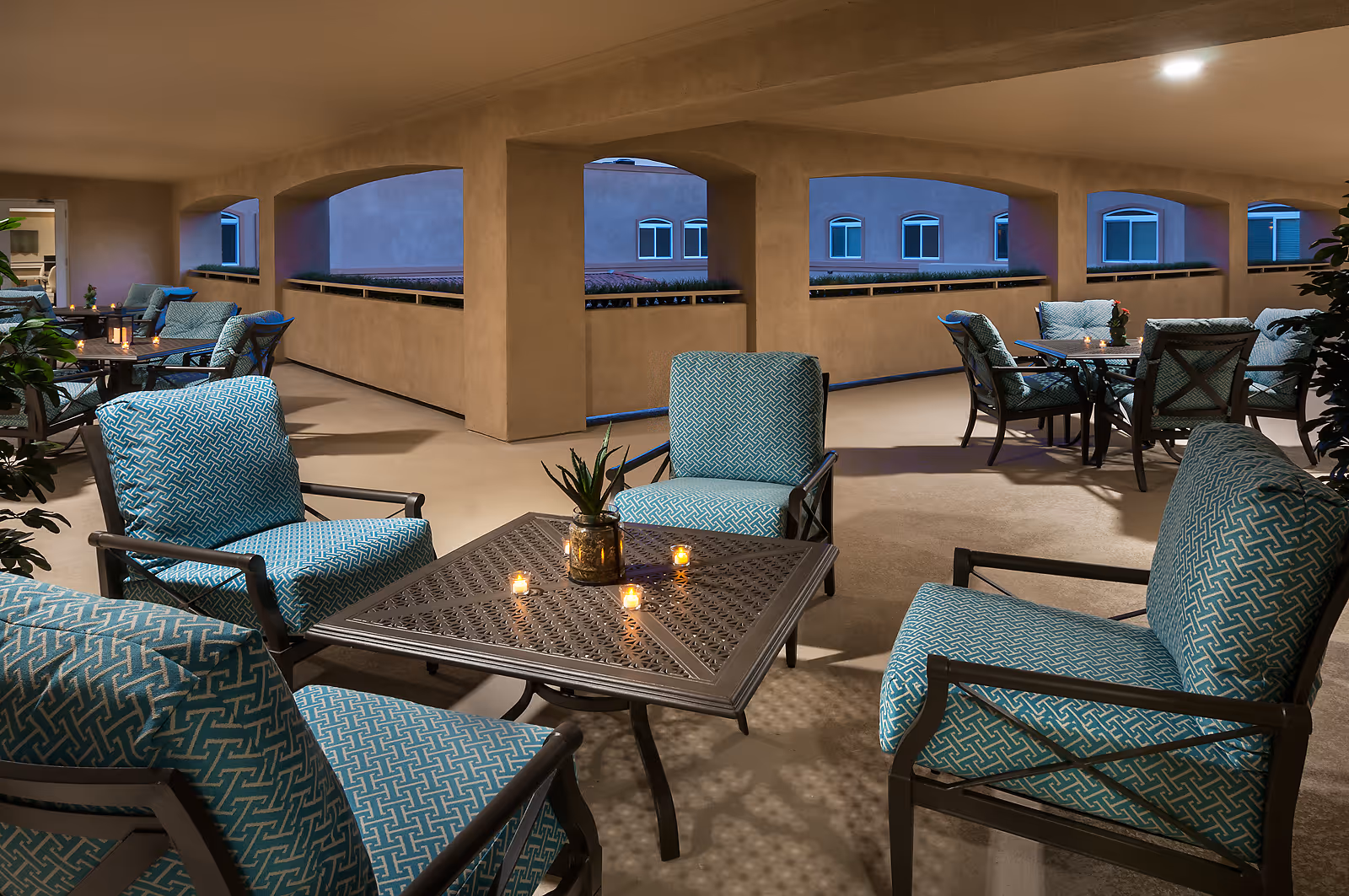Covered outdoor seating area with cushioned chairs arranged around metal tables. Small candles and a potted plant are on the tables. The space has arched openings showing a view of windows on an adjacent building.