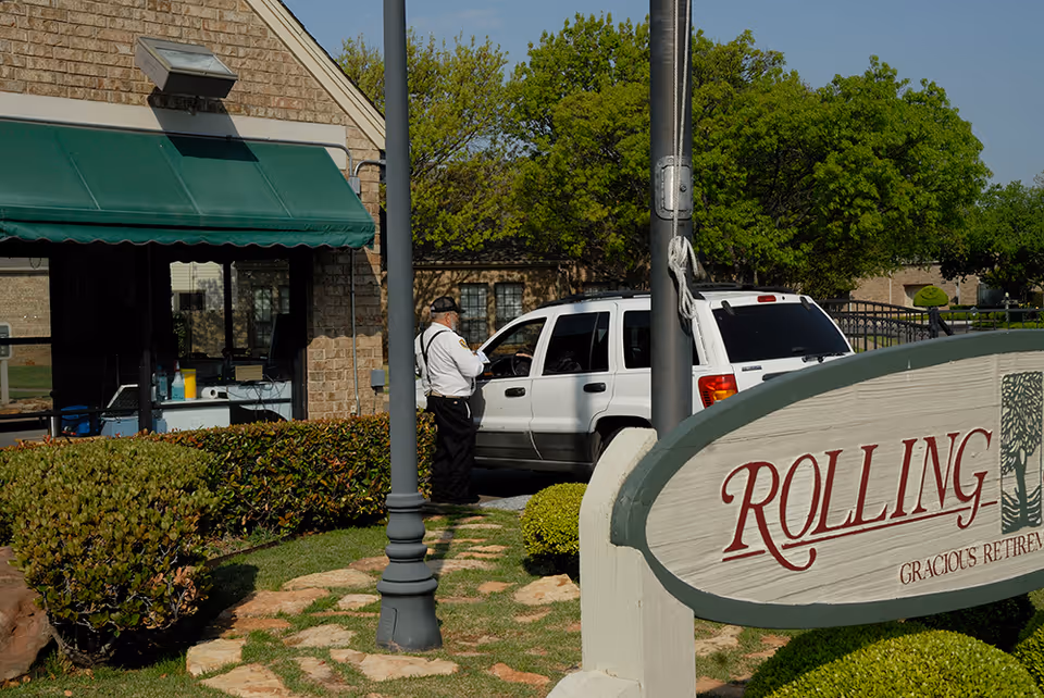 A security guard stands next to a white SUV parked near a building with a green awning. In the foreground, there is a sign that reads 'ROLLING' with some additional text partially visible. The area is landscaped with bushes and trees under a clear sky.