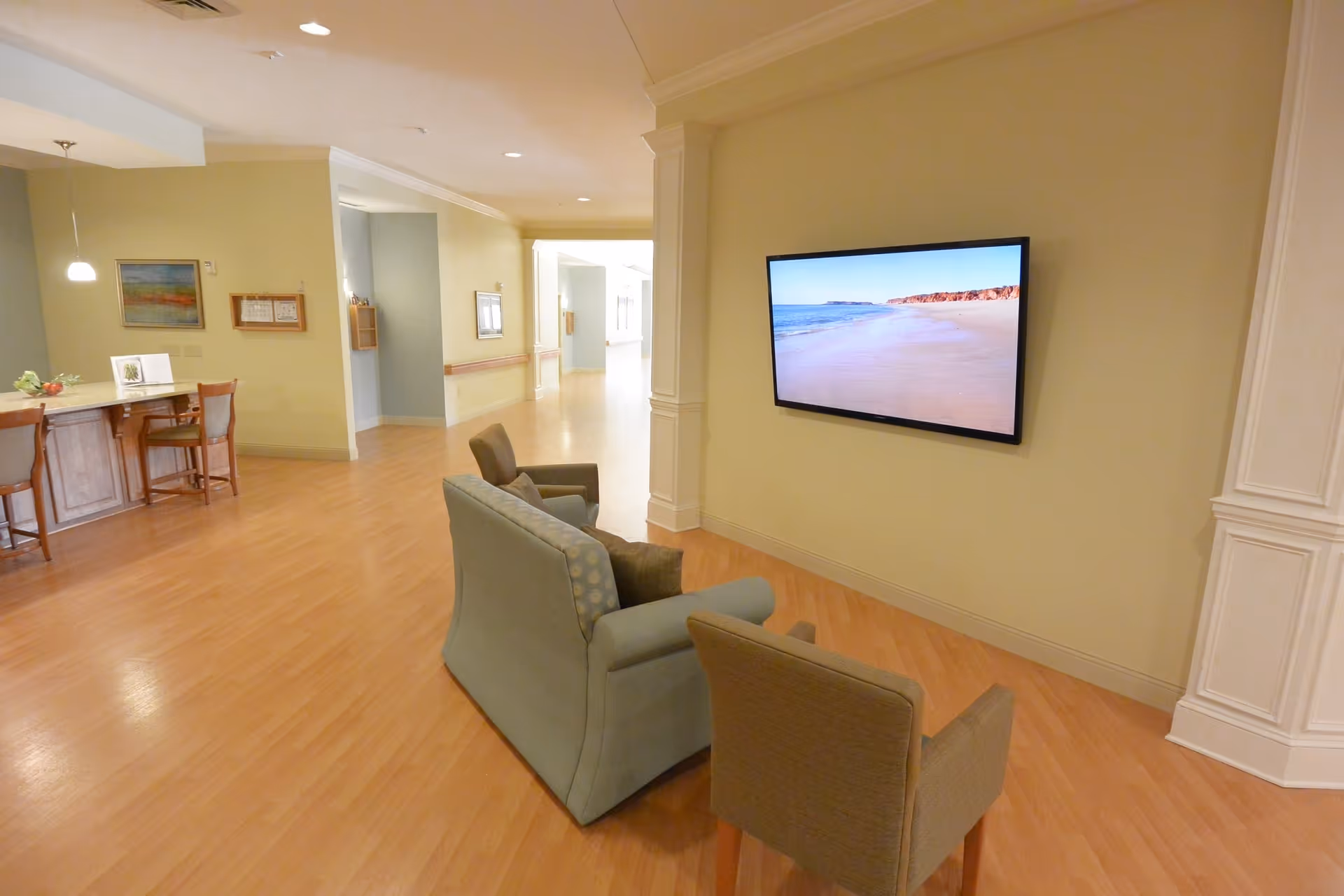 A cozy seating area in a senior living facility with a sofa and two armchairs facing a wall-mounted flat screen TV displaying a beach scene. The room has light wood flooring, soft yellow walls, and a hallway leading to other areas. A dining area with chairs and a table is visible in the background.