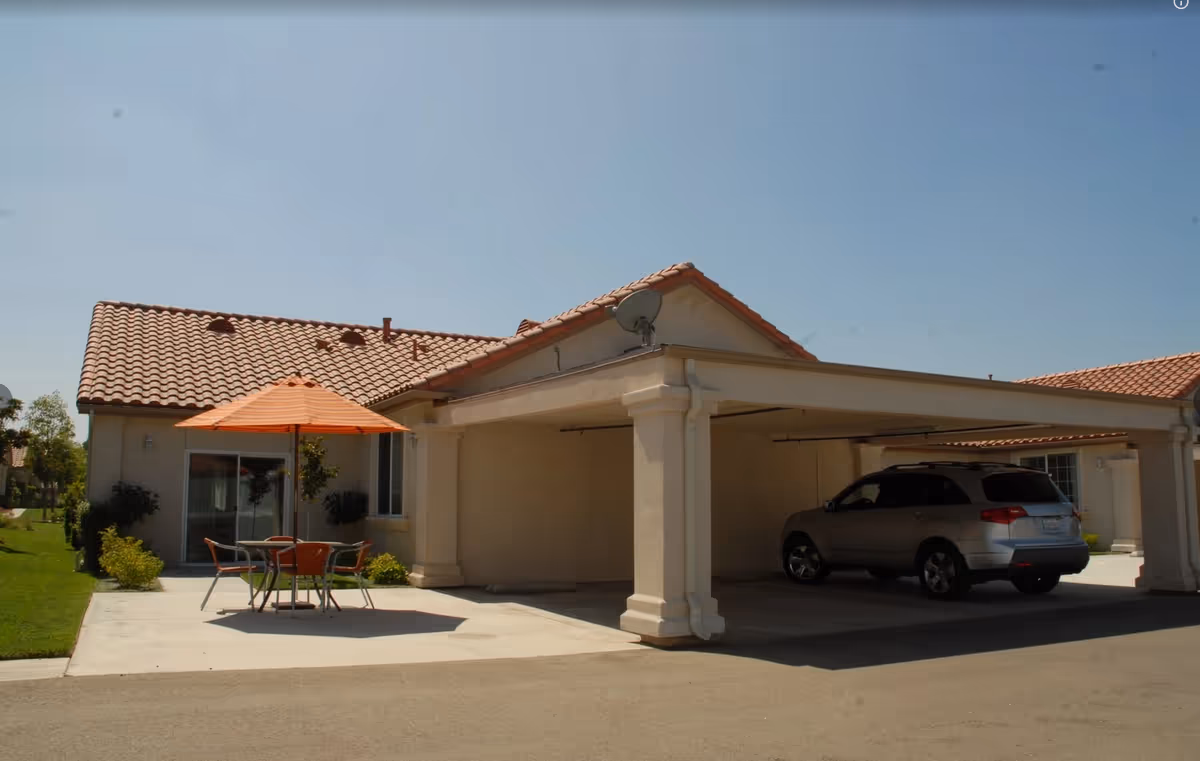 Exterior front of a single-story retirement home with a tiled roof, covered carport with a parked SUV, and a patio table with an orange umbrella.