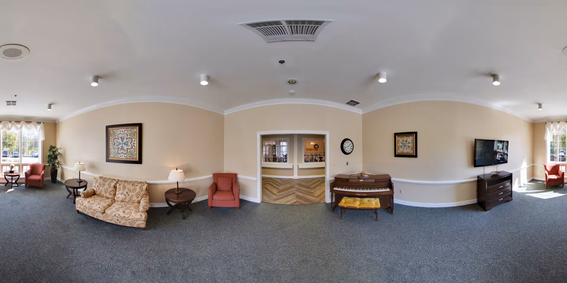 Panoramic view of a bright senior living common room with sofas, armchairs, a piano, a TV, and large windows.