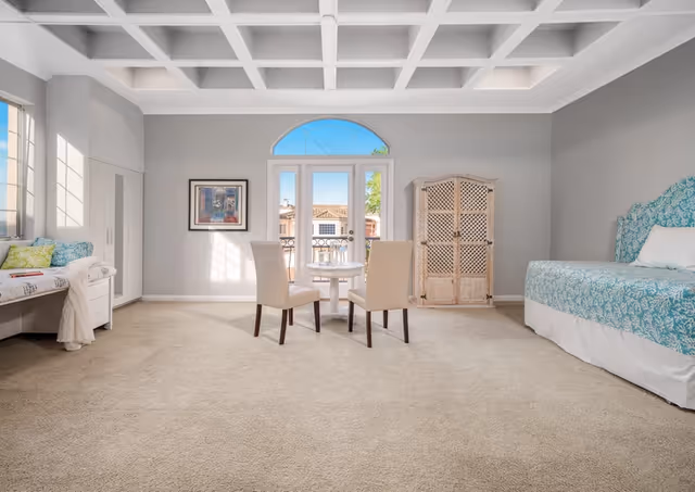 A spacious, well-lit room with beige carpet and light gray walls. The room features a bed with a blue and white patterned headboard and bedding on the right, a small round table with two beige chairs in the center, a white cabinet with lattice doors against the back wall, and a window seat with cushions and a throw blanket on the left. There is a large window and a glass door with an arched transom letting in natural light and showing a view of rooftops outside. The ceiling has a white coffered design.