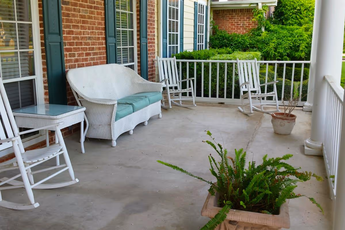 A covered outdoor porch area with white wicker furniture including a loveseat with green cushions, a glass-top side table, and several white rocking chairs. There are potted plants on the concrete floor and green bushes along the railing. The porch is attached to a brick building with windows and green shutters.