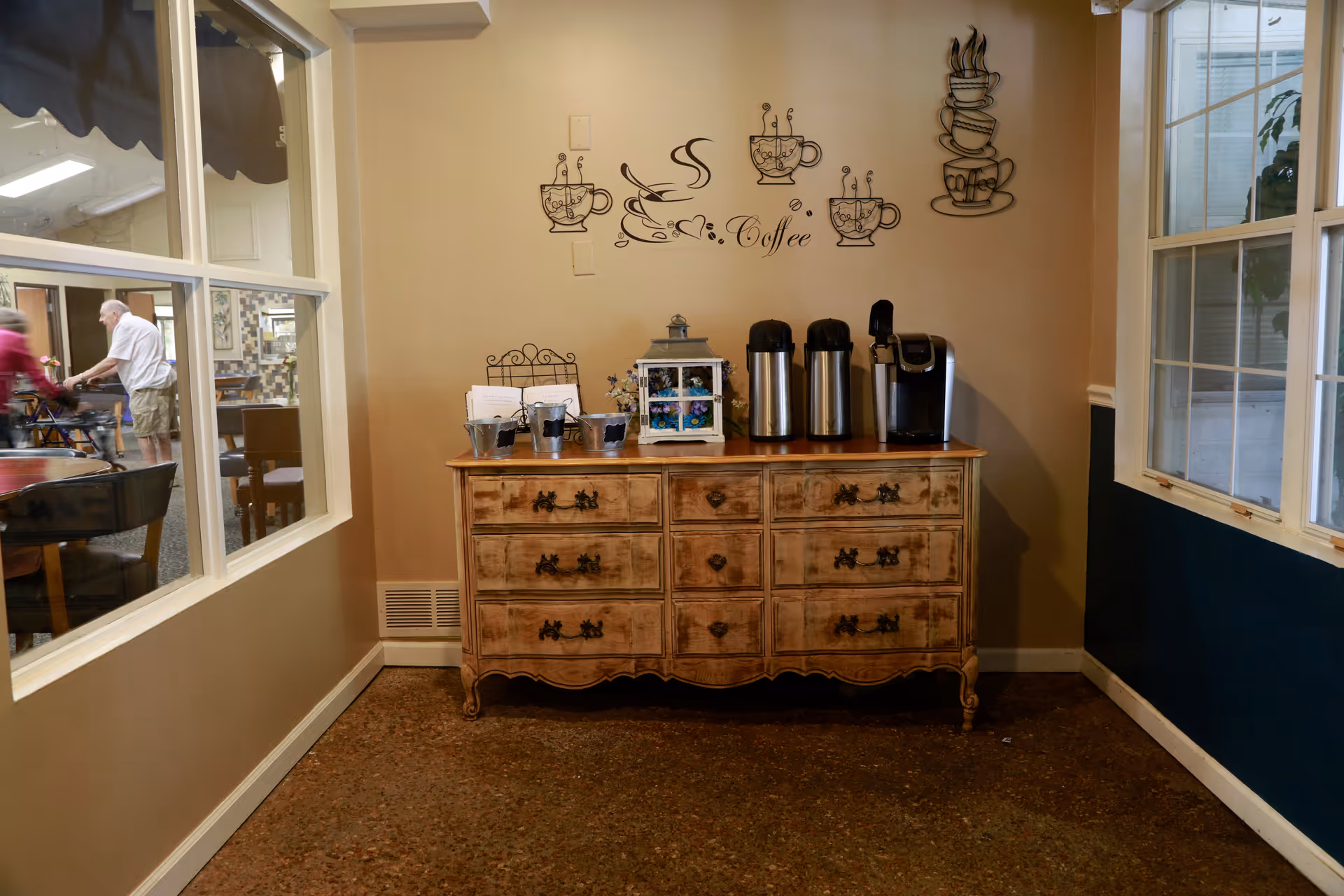 A vintage wooden sideboard set up as a coffee station with airpots and a single-serve brewer under coffee-themed wall decals in a common area.