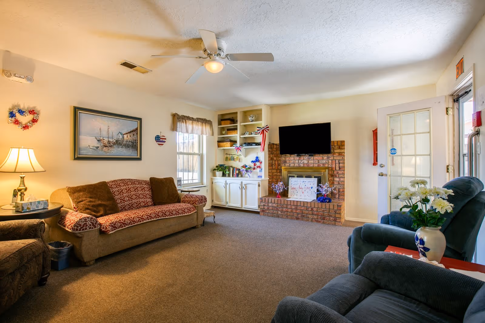 A cozy living room in an assisted living facility with a beige couch covered with red patterned throws and brown pillows, a side table with a lamp, a window with a valance, built-in white shelves and cabinets decorated with patriotic items, a brick fireplace with a mounted flat-screen TV above it, and two blue armchairs with a vase of white flowers on a small table. The room has beige walls, carpeted floor, and a ceiling fan with a light.