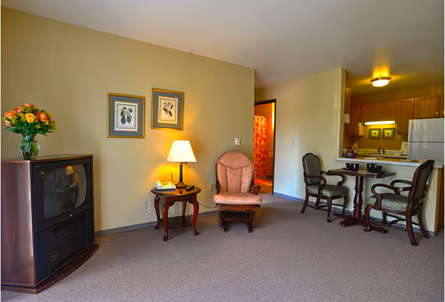 A cozy living area in a senior living facility with a vintage television on a wooden stand, a side table with a lamp and telephone, an upholstered armchair, and a small dining table with two chairs. The kitchen area with wooden cabinets and a refrigerator is visible in the background, along with a doorway leading to another room with a red patterned curtain.