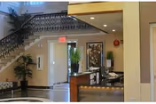 Interior view of a facility lobby or reception area with a staircase featuring decorative black railings, potted plants, a reception desk with computer monitors, and framed artwork on the walls.