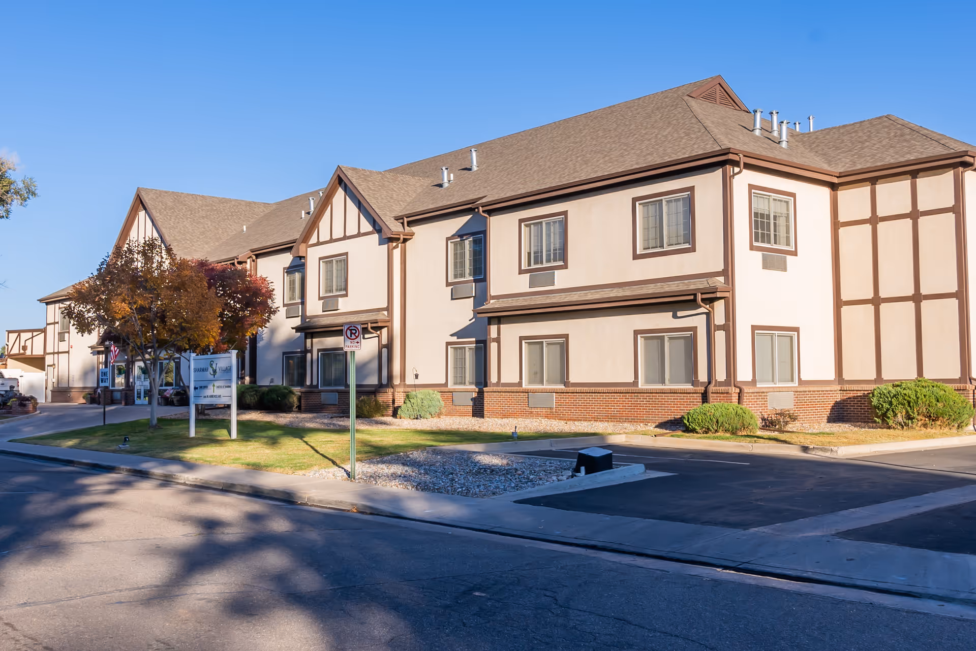 Two-story beige and brown Tudor-style assisted living building front with a parking area, small lawn, and signage under a clear blue sky.