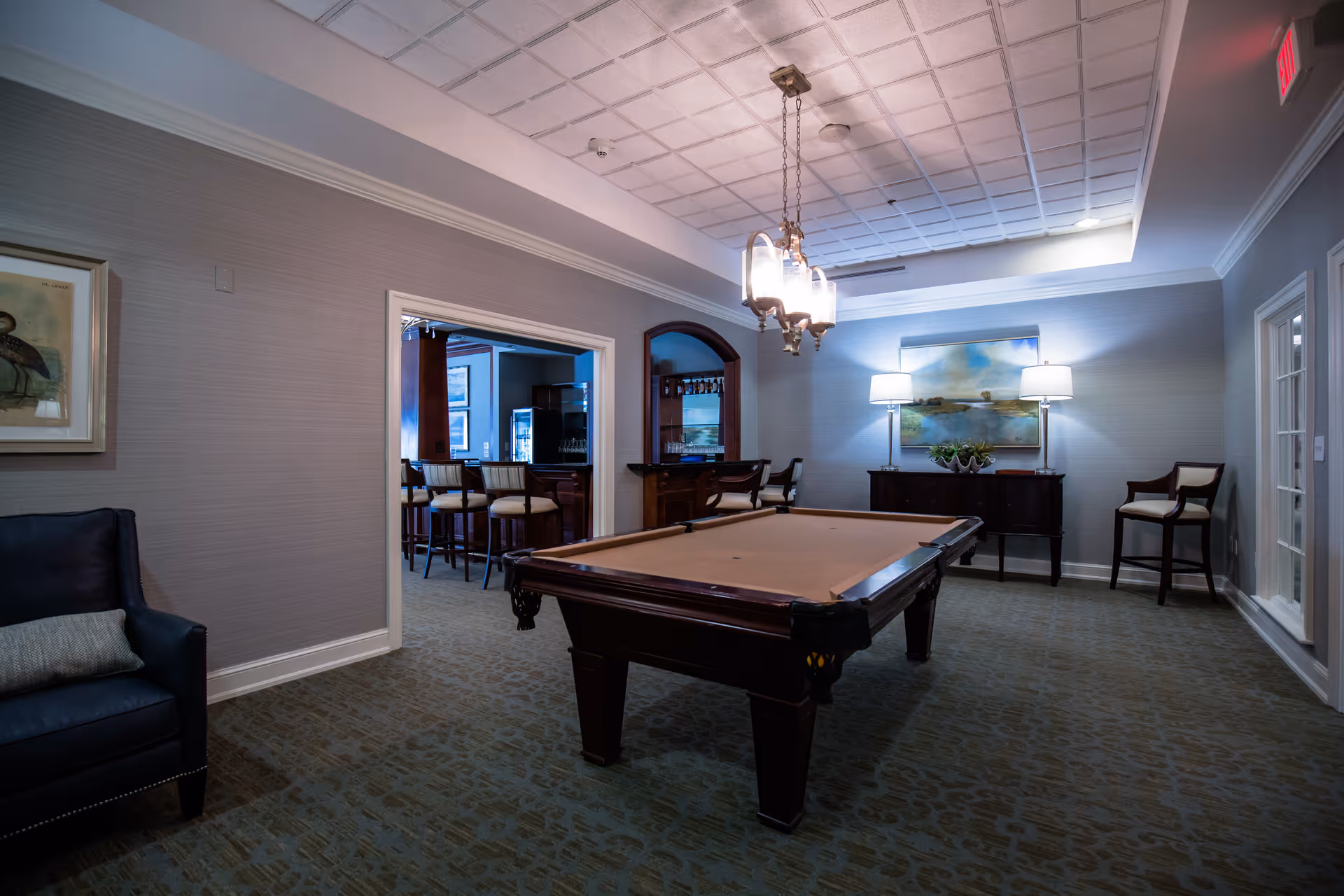A well-lit game room featuring a pool table in the center, a dark blue armchair with a cushion on the left, and a bar area with stools visible through an open doorway. The room has a patterned carpet, a chandelier hanging above the pool table, and a sideboard with two lamps and a landscape painting on the far wall.
