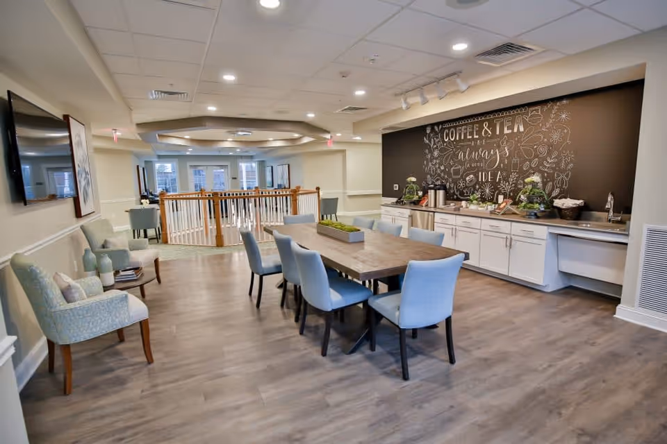 A bright and spacious common area with a long wooden table surrounded by blue upholstered chairs. On the right side, there is a countertop with white cabinets underneath and a black chalkboard wall behind it featuring decorative writing that says 'Coffee & Tea always a good idea' along with various drawings. The floor is wood, and there are additional seating areas with armchairs on the left side. The ceiling has recessed lighting and a decorative tray ceiling in the center.