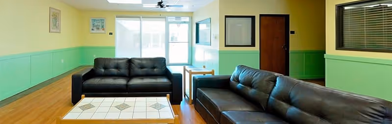 A bright and clean common area with two black leather sofas facing each other, a tiled coffee table in between, light green and yellow walls, wooden flooring, a ceiling fan, and a glass door leading outside.