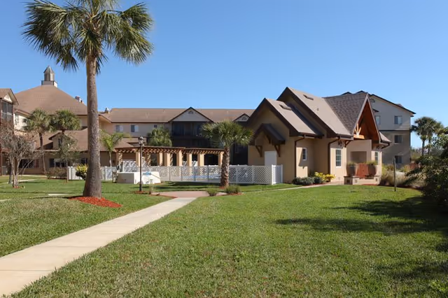 A well-maintained outdoor area of a senior living facility with a paved walkway leading through a green lawn, palm trees, and a building with a beige exterior and brown roof in the background under a clear blue sky.