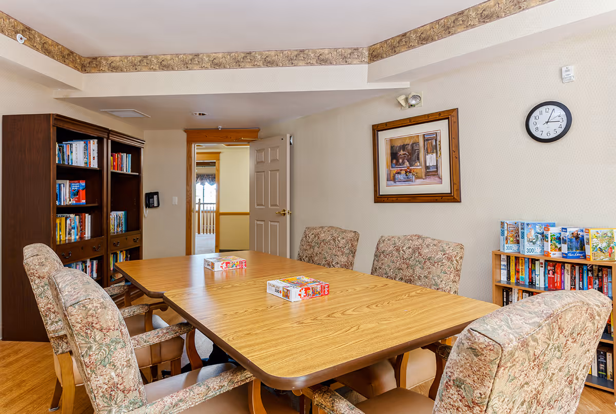 A cozy room with a wooden table surrounded by six cushioned chairs with floral upholstery. On the table are two puzzle boxes. The room has two bookshelves filled with books and puzzles. A framed painting hangs on the wall above the chairs, and a clock shows the time as 2:10. The door is open, revealing a hallway with natural light.