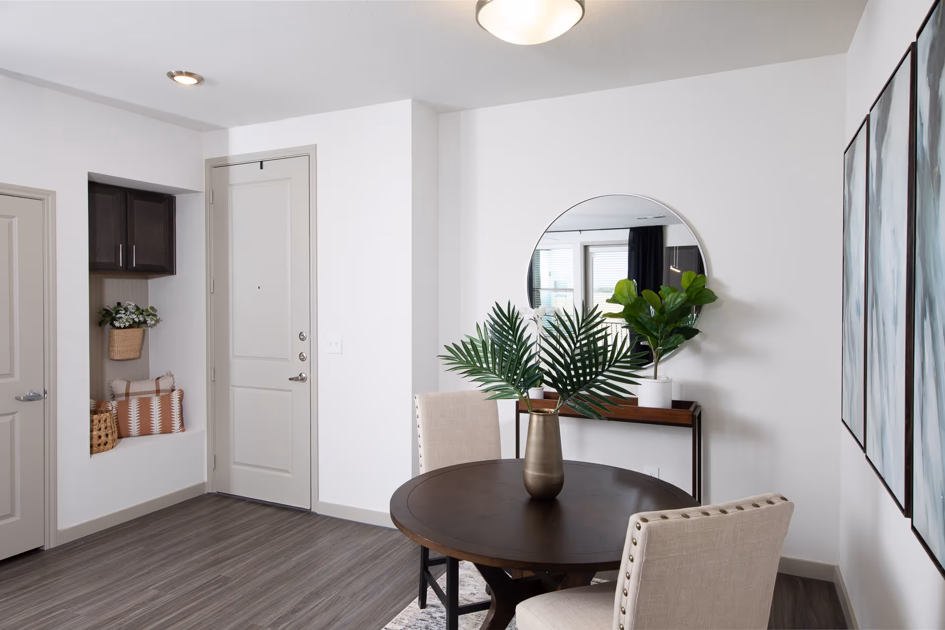 A modern dining area with a round dark wood table and two beige upholstered chairs. A bronze vase with green palm leaves is centered on the table. Behind the table is a console table with a round mirror above it and a potted plant. To the left is a white door, a small built-in nook with cushions and a basket, and a cabinet above it. The floor is a dark wood laminate and the walls are white.