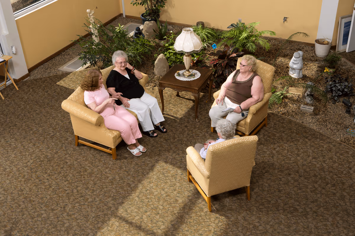 Four elderly women sitting and chatting in a cozy indoor lounge area with beige armchairs arranged around a wooden side table with a decorative lamp. Behind them is an indoor garden with various plants and small statues, set against a yellow wall.