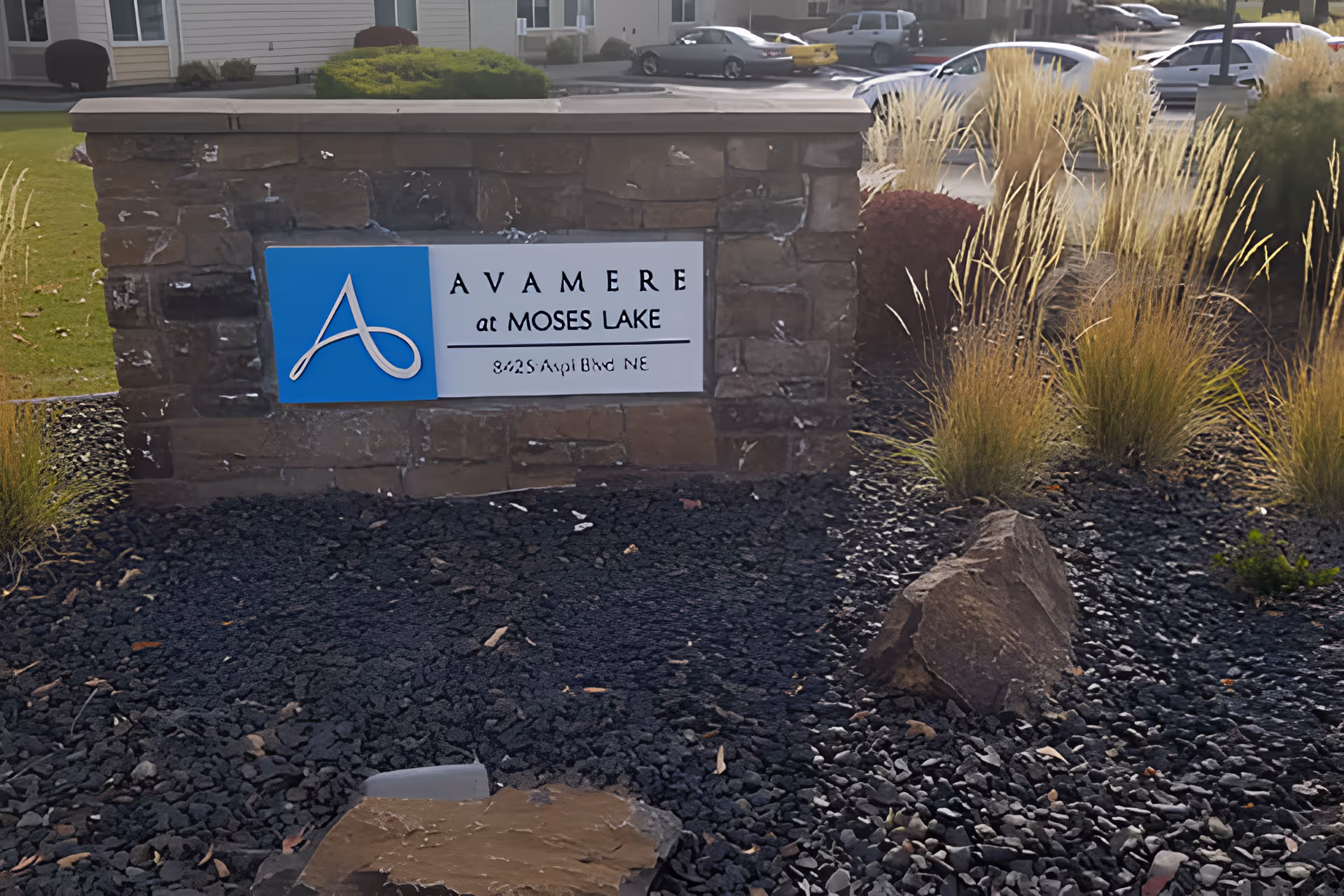 Stone sign with a blue and white plaque displaying the logo and name 'Avamere at Moses Lake' along with the address '8425 Aspi Blvd NE', surrounded by landscaping with black gravel, rocks, ornamental grasses, and bushes, with parked cars and part of a building visible in the background.