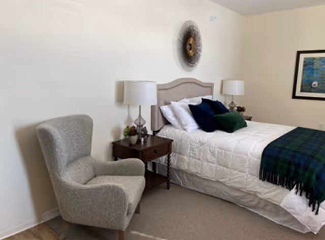 Neatly made bed with several pillows and a plaid throw, flanked by nightstands and lamps, with an upholstered armchair and wall art in a bedroom.
