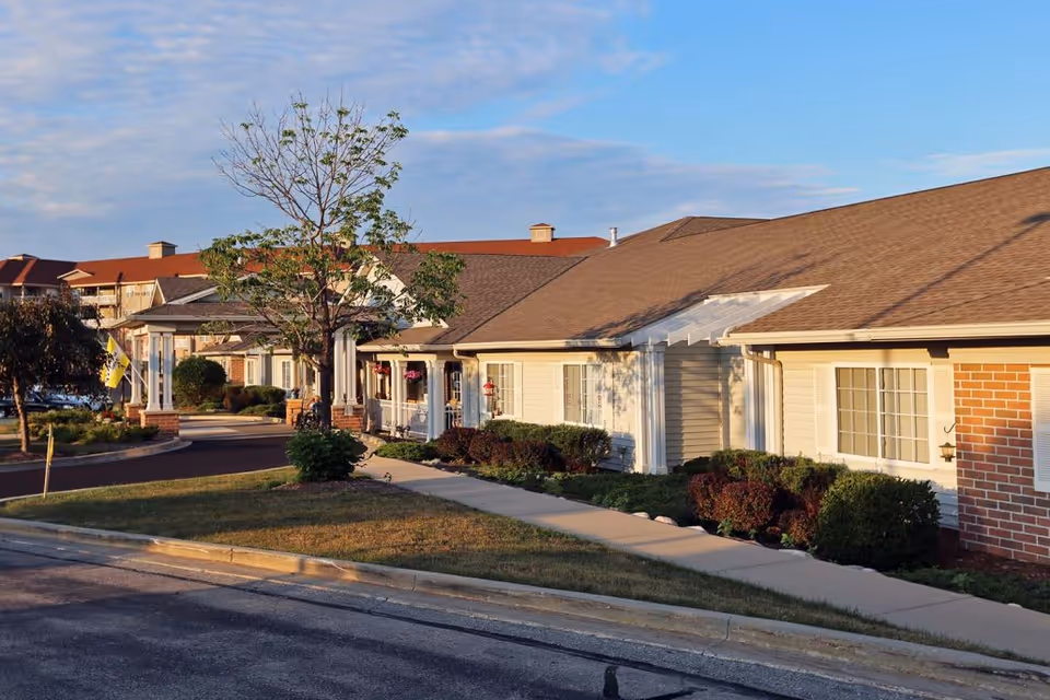 Exterior view of Brenwood Park Assisted Living facility showing a single-story building with beige siding, white trim, and a brown shingled roof. There is a sidewalk and landscaped bushes in front, with a tree and a covered entrance visible. The sky is partly cloudy with soft sunlight illuminating the scene.