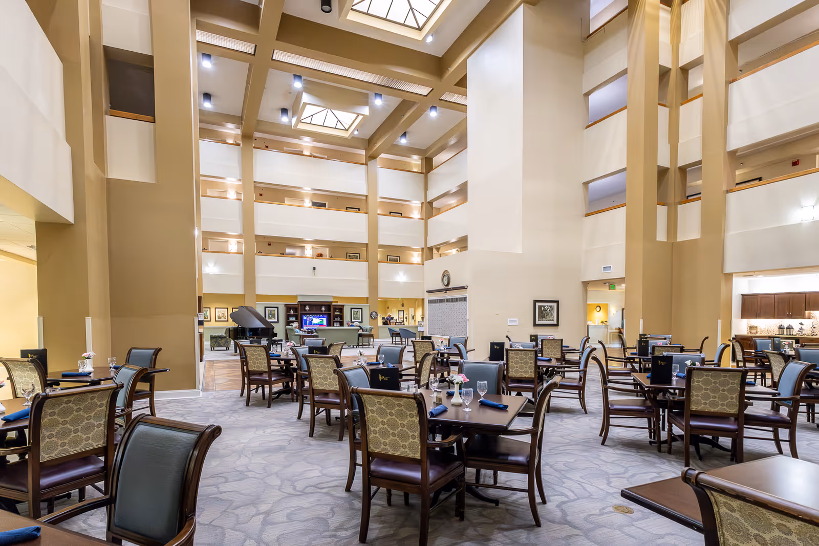 Spacious dining area in Elison Assisted Living of Bella Vita with multiple tables and chairs arranged neatly. The room features high ceilings with skylights, warm lighting, and a grand piano in the background near a seating area with a television and framed artwork on the walls.
