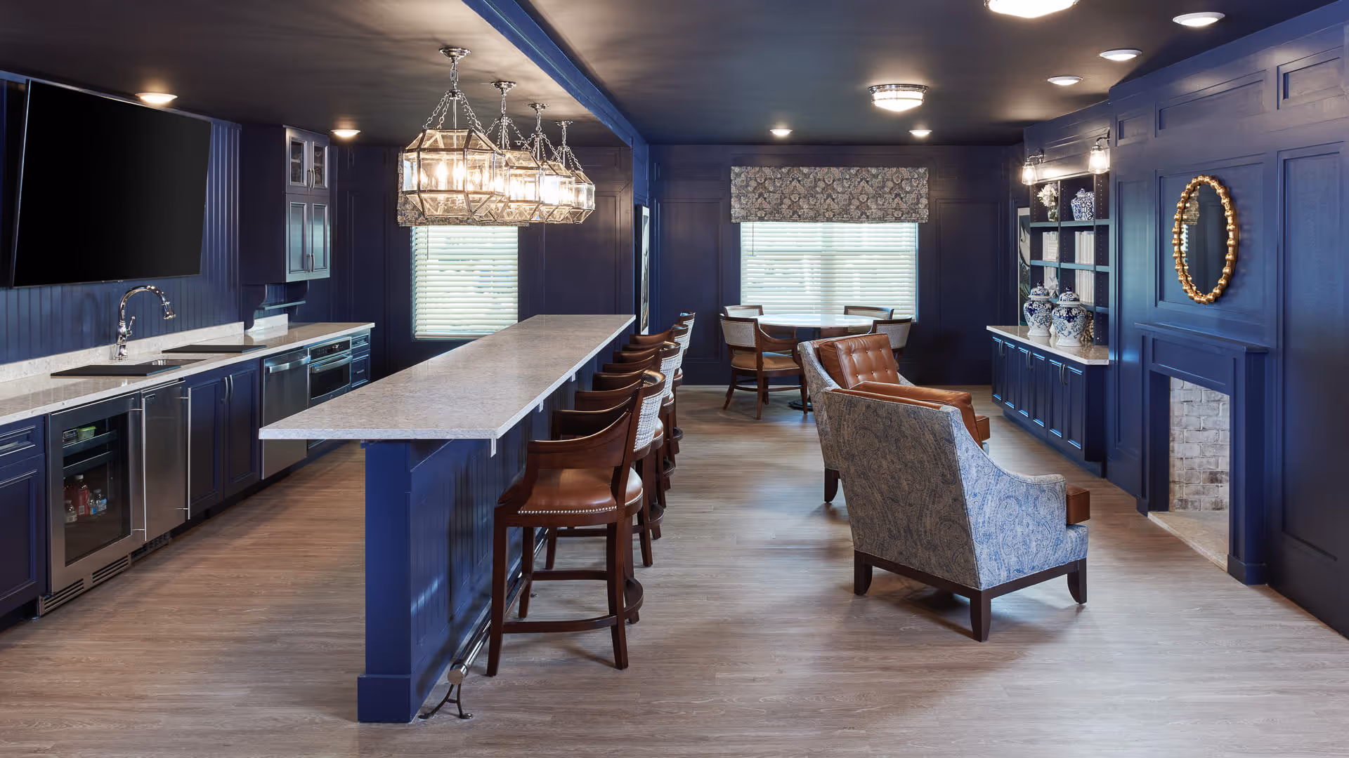 A spacious interior room with dark blue walls and cabinetry, featuring a long kitchen island with bar stools, a sink, and appliances on the left side. The room also includes a seating area with two armchairs facing a fireplace with a round mirror above it, and a small dining table with chairs near a window with blinds and a patterned valance.